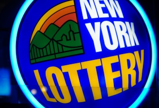 A bright, circular neon sign with a blue background, featuring the words 'New York Lottery' in bold white and yellow letters. The sign includes a colorful illustration of a landscape with green mountains, an orange-red sunset, and a bridge, all encapsulated within the circle.