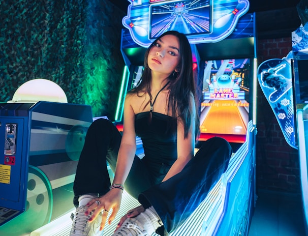 A person sitting on an arcade game machine platform with neon lights in the background. The arcade game is called 'Lane Master' and features a colorful design with bowling-themed imagery.