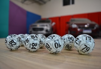 A collection of white lottery balls with black numerals scattered on a wooden surface. Two vehicles are blurred in the background, parked in a room with multicolored walls including teal, purple, blue, and red panels.