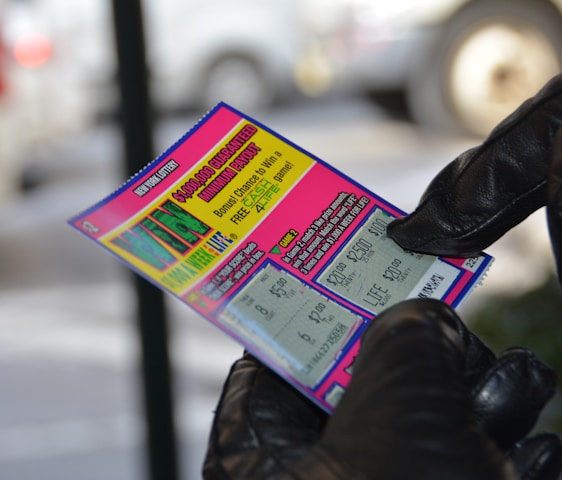 A person wearing black gloves is holding a colorful lottery scratch-off ticket. The ticket includes information about a $1,000,000 guaranteed minimum payout and features multiple games with various prize amounts.
