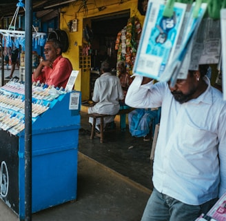 A street vendor stall displaying an array of lottery tickets clipped to a bright blue kiosk. A man in a red shirt leans on the counter, appearing thoughtful, while another man in a white shirt adjusts his hat and walks by. A variety of products hangs from strings above, and a colorful display fills the background with vibrant signs and packets. The scene suggests a busy, lively marketplace.